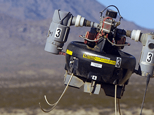 A small, black, unmanned aerial vehicle (UAV) with a yellow label and a white number 3 on its side, flying in a desert landscape.