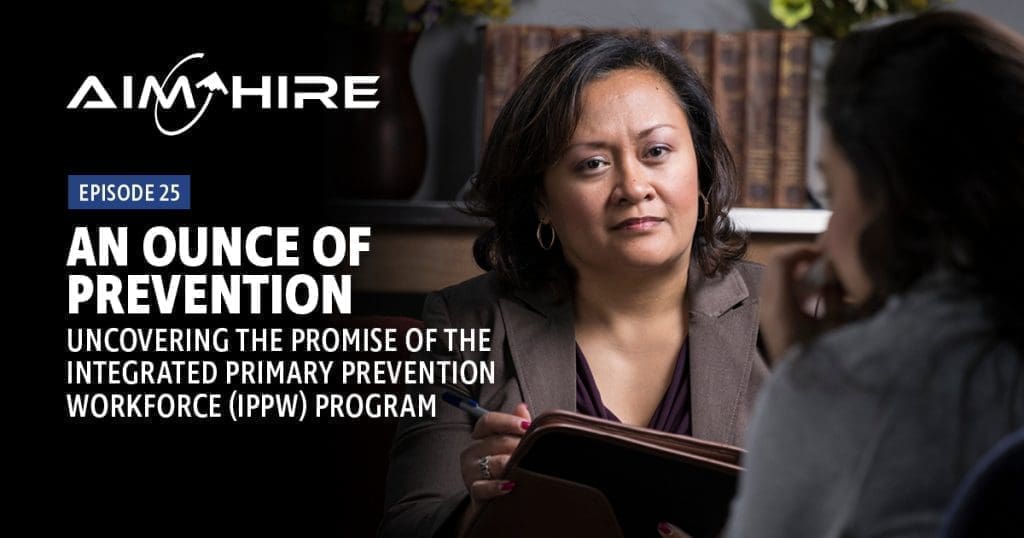 Aim Hire Episode 25: A woman in a brown blazer holding a tablet, with a person in the foreground and a bookshelf in the background.
